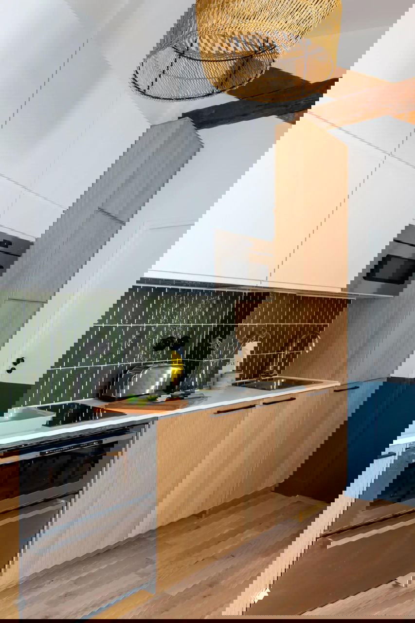 Кухонная зона: нижние фасады из светлого дерева, белые высокие шкафы, тёмно‑зеленая плитка фартука и ротанговая подвесная лампа над рабочей поверхностью.