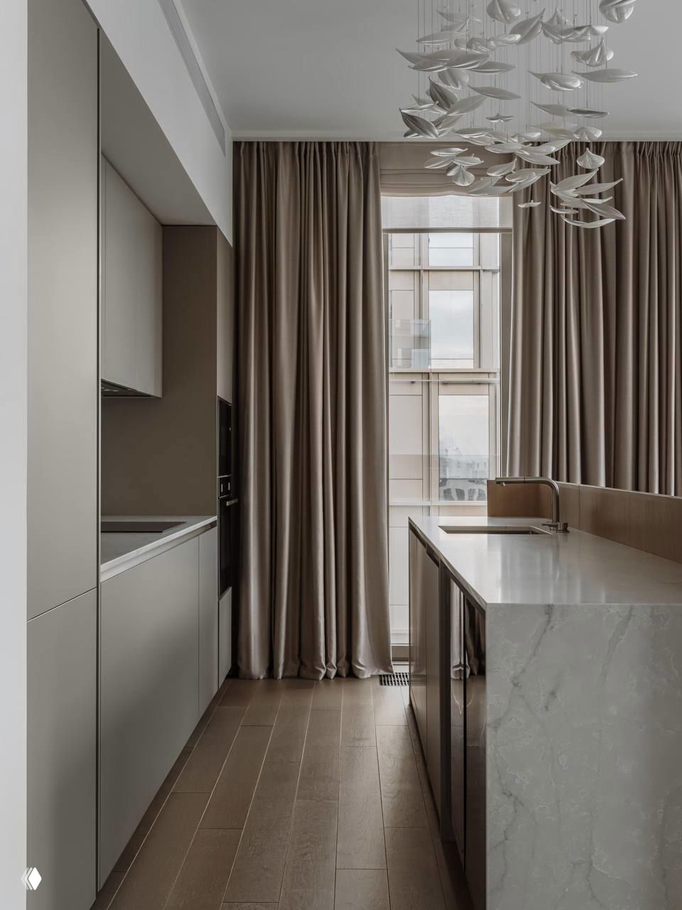Сдержанная современная кухня в бежево-коричневой гамме с мраморным островом, панорамным окном и удобной проходной зоной между рабочими рядами.