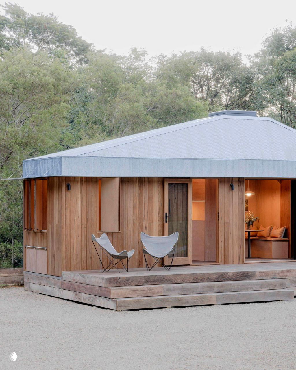 Общий вид дома на террасе: деревянное крыльцо, пара лёгких кресел и компактная архитектура, вписанная в зелёный пейзаж.