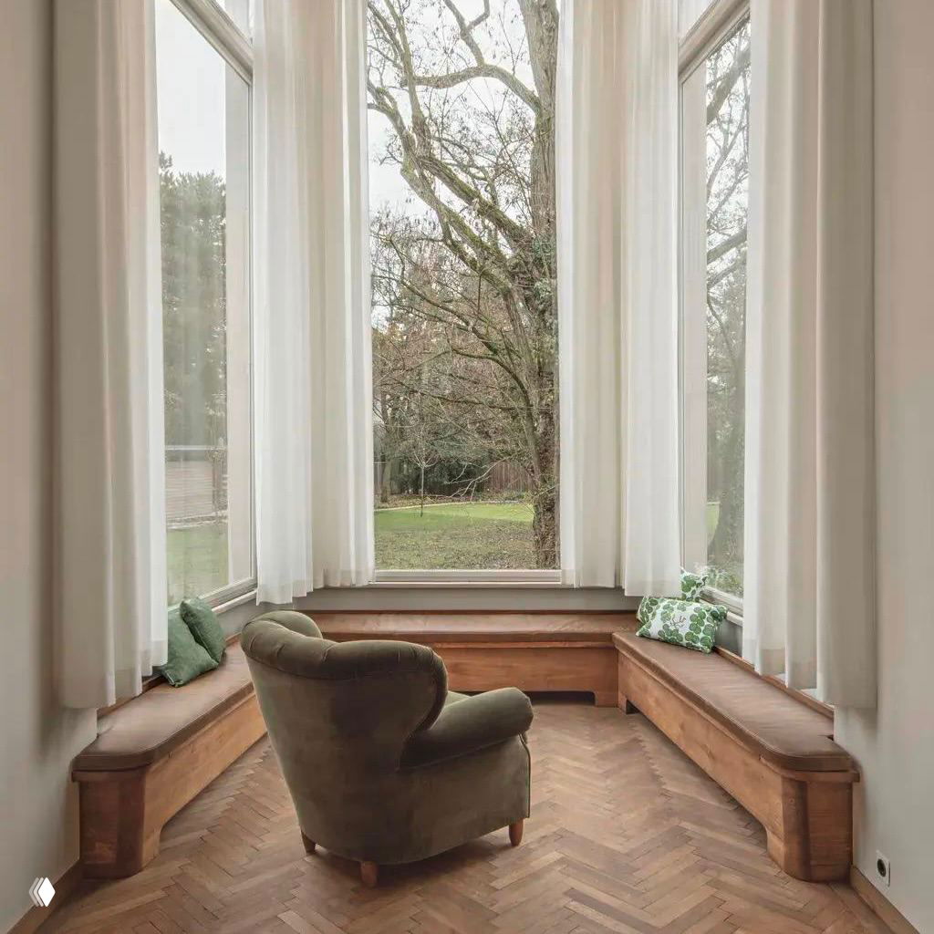 Уютный эркер с большим панорамным окном в вилле Beer: скамьи вдоль окон и кресло, вид на сад через высокие световые проёмы и лёгкие шторы.