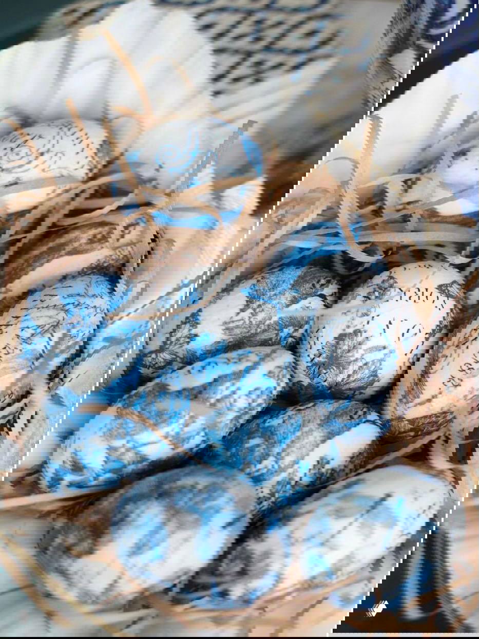 Корзинка с пасхальными яйцами в технике декупажа: синие цветочные узоры и текстовые фрагменты на белом фоне, декоративная стружка вокруг.