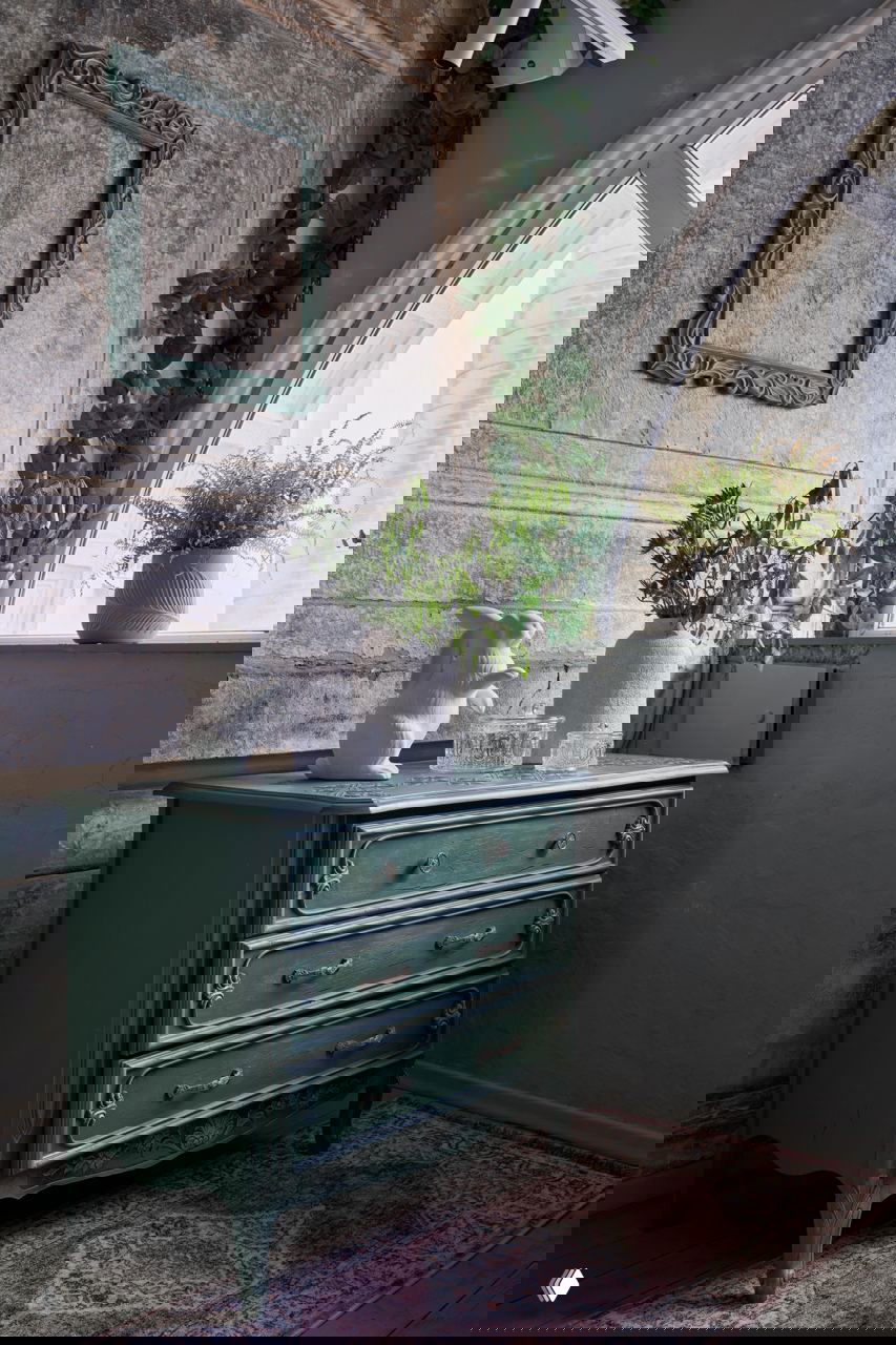 Деталь интерьера: зелёный винтажный комод у арочного окна, вазы с растениями и декоративная фигурка на фоне патинированной стены и окна.