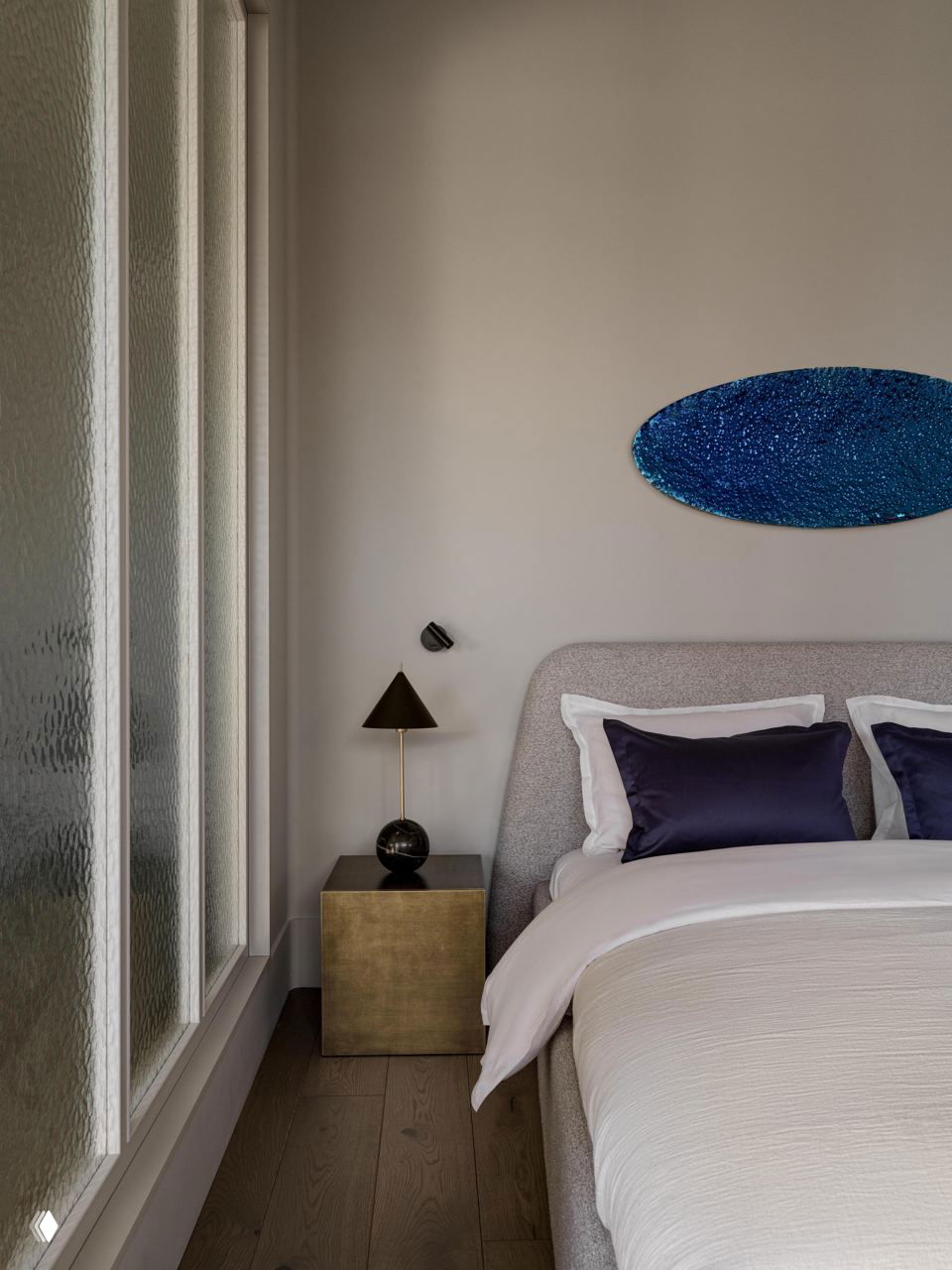 Спальня с мягкой обивкой кровати, тёмно-синими подушками и декоративным настенным арт-объектом; спокойная палитра и натуральные материалы создают уют.