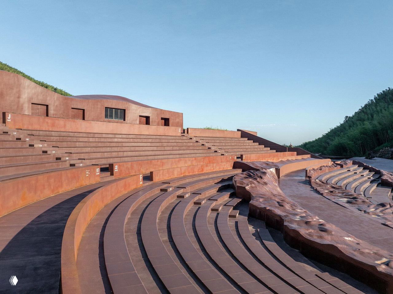 Вид с уровня зрительских мест на ступенчатые ряды амфитеатра и волнообразные ограждающие стены на фоне зелёных холмов и ясного неба.