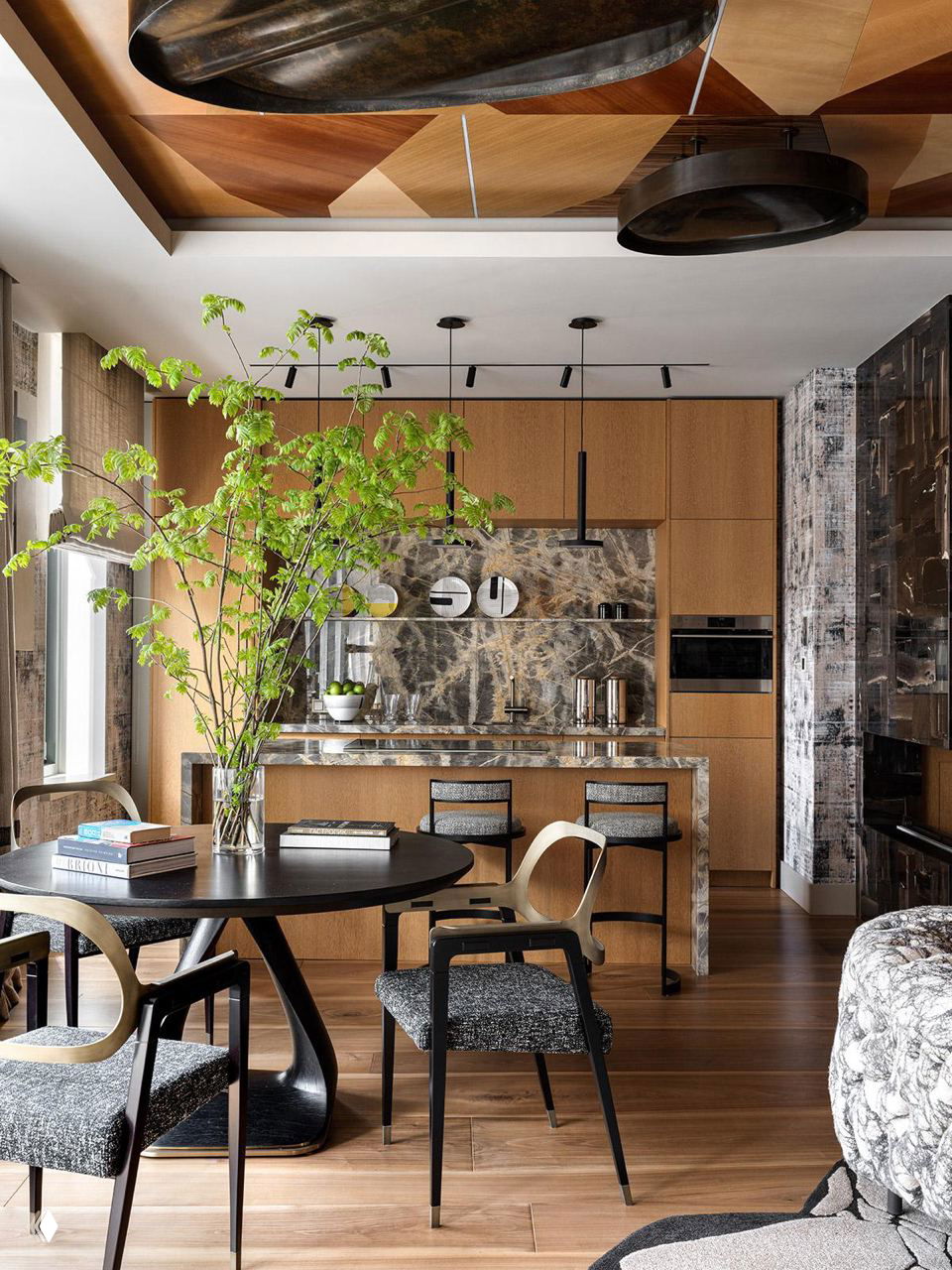 Столовая и кухня в едином пространстве: круглый стол, стулья, остров с мраморной столешницей и крупный напольный рисунок на деревянном полу при естественном свете.