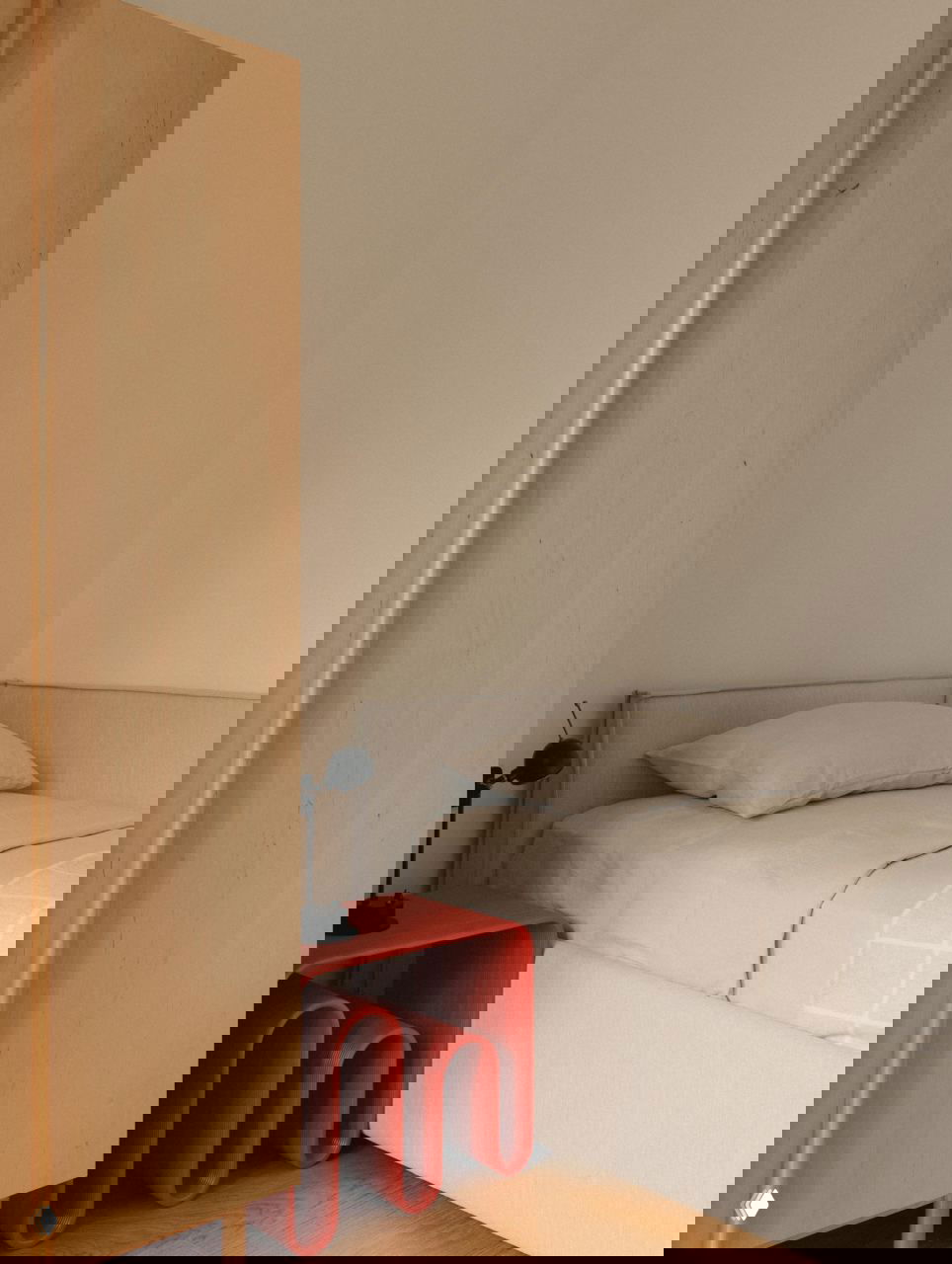 Небольшая соседняя комната: односпальная кровать, яркий фигурный красный прикроватный столик и светлый деревянный шкаф в лаконичном минималистичном решении.