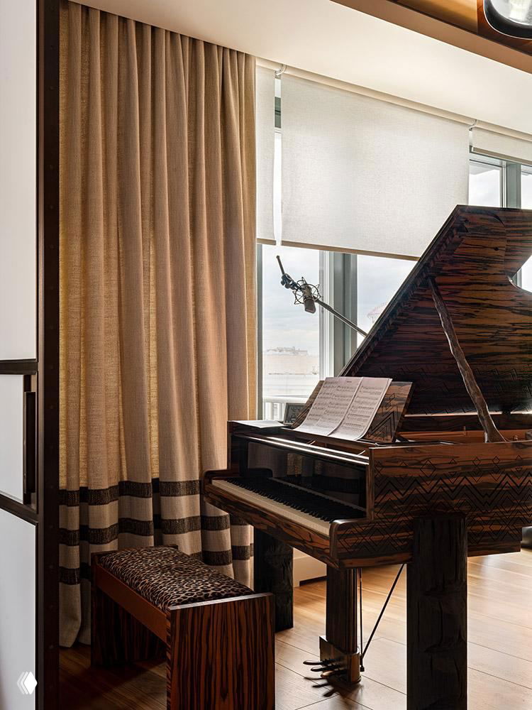 Крупный план рояля у окна: рояль в резной деревянной отделке, нотная тетрадь, микрофон и мягкие шторы на фоне городской панорамы за окном.