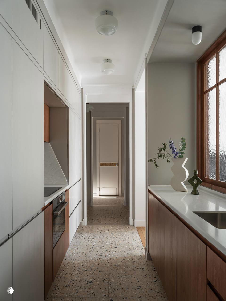 Узкая проходная кухня с белыми верхами и деревянными нижними шкафами, терраццо-пол и большое окно создают ощущение глубины и света в вытянутом пространстве.