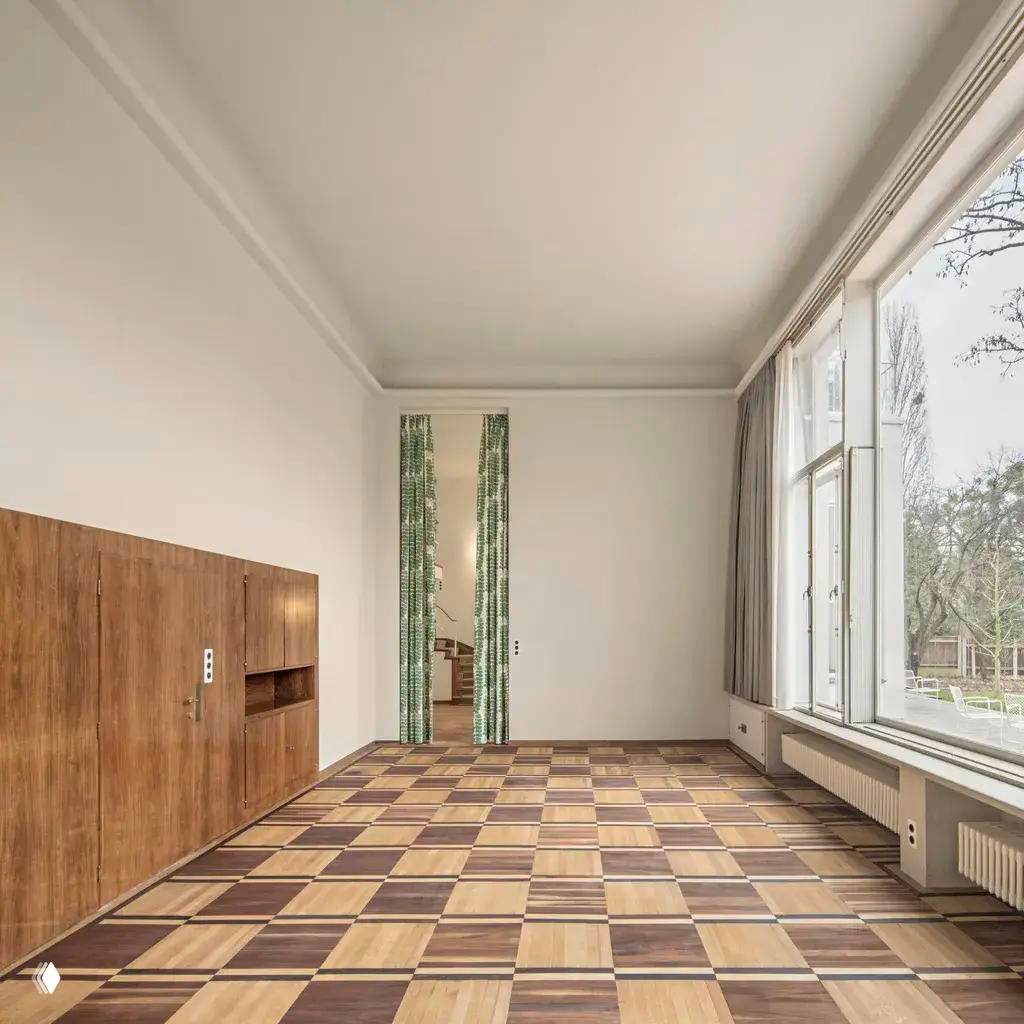 Просторная комната виллы Beer с паркетообразным рисунком пола и большими окнами: светлые стены и встроенные хранилища в духе 1930-х годов.