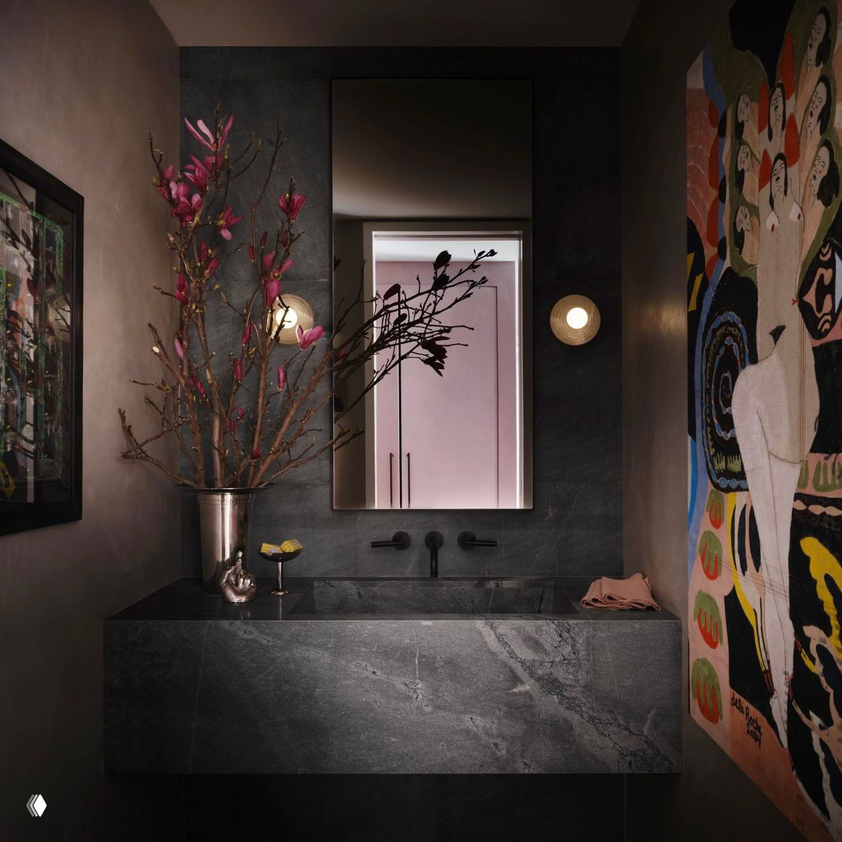 Небольшая мраморная раковина в тёмной ванной, украшенная цветочной веткой и яркой живописной работой на стене — сочетание строгого камня и экспрессивного искусства.