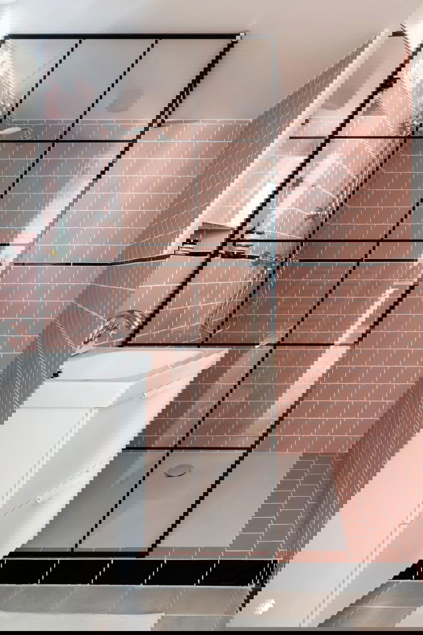 Ванная комната с розовой плиткой метро, чёрной металлической стеклянной перегородкой в стиле лофт и компактной белой тумбой у раковины в небольшом пространстве.