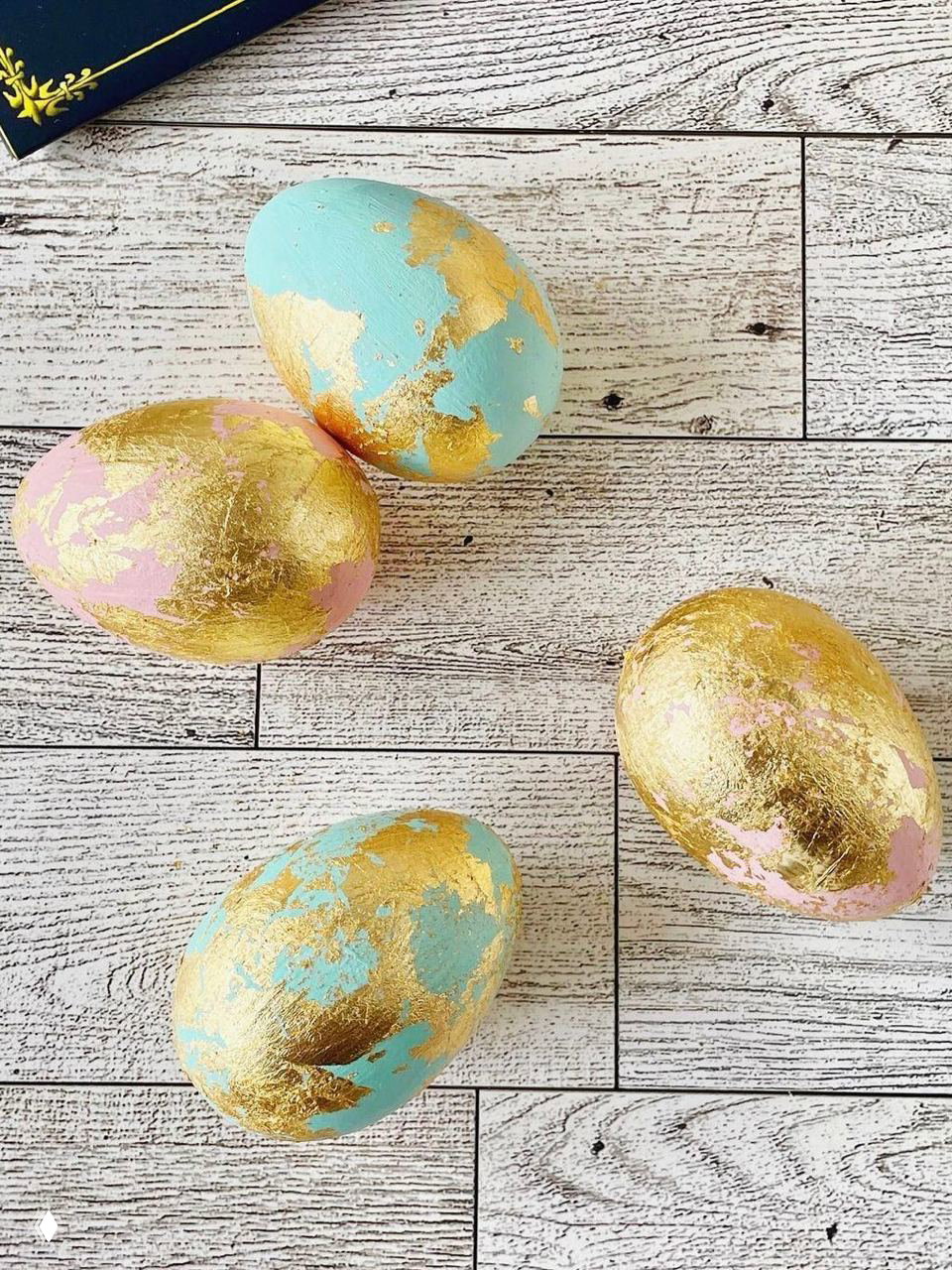 Пасхальные яйца, украшенные золотой сусалью: бирюзовая и розовая основа с наложением золотых листов, расположены на светлой деревянной поверхности.