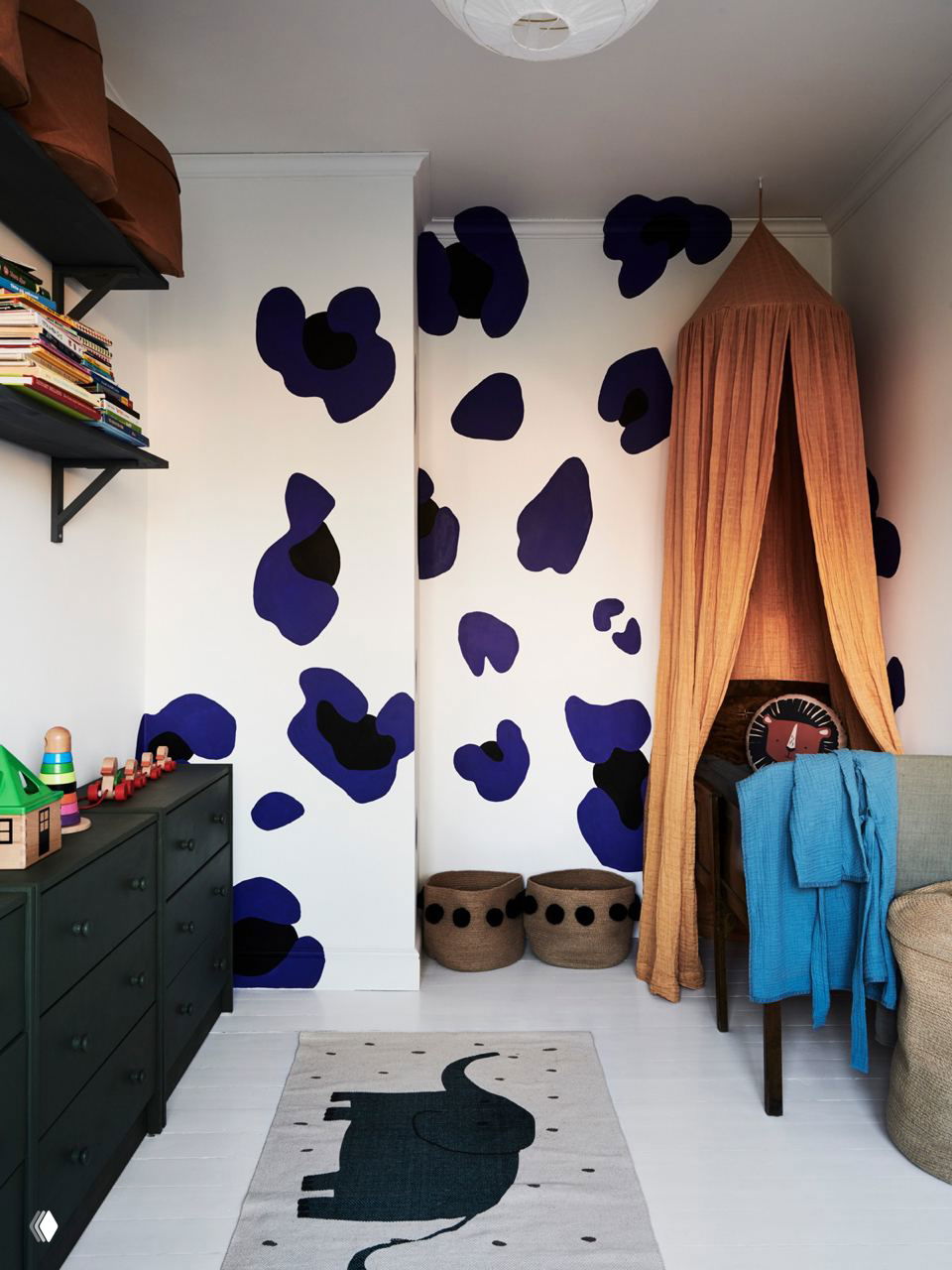 Яркий уголок детской: оранжевый балдахин-«шалаш» у стены с графичными синими пятнами, детские игрушки и мебель в контрастных цветах.