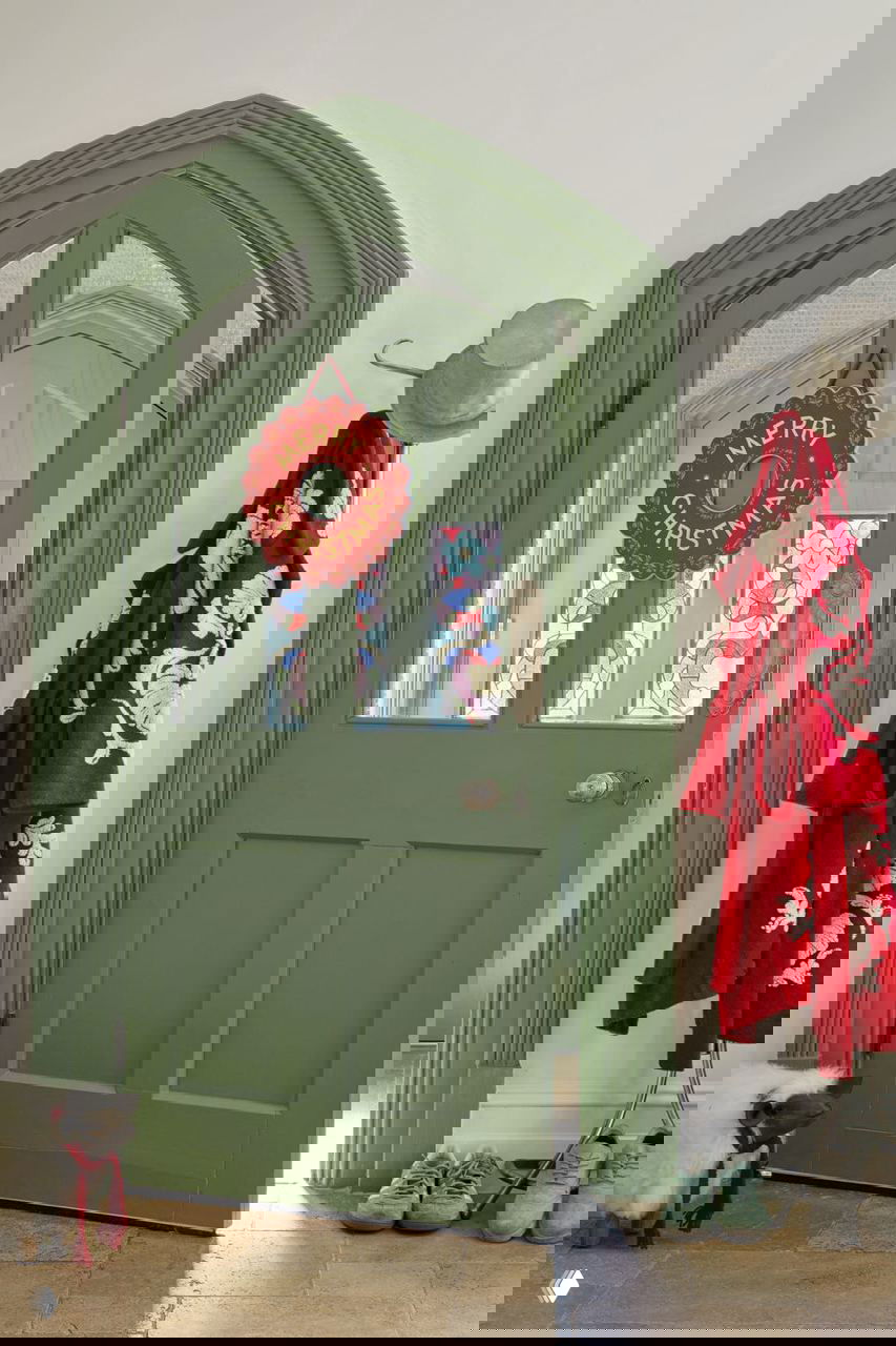 Зеленая входная дверь с рождественской подвеской «Merry Christmas», у пола — обувь и уютные детали в прихожей английского дома Холли Такер.