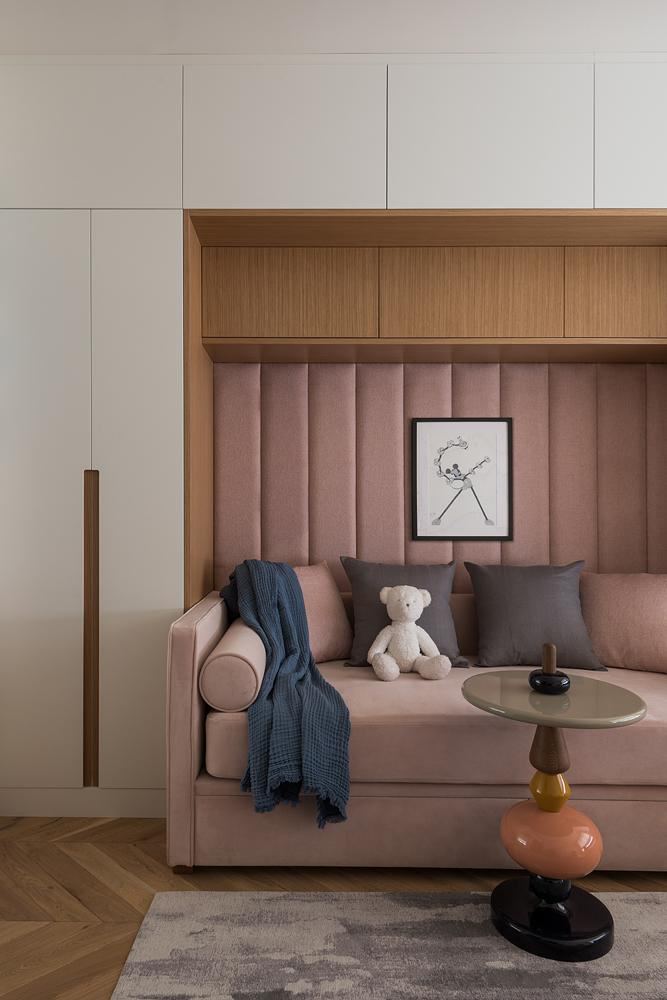 Уютная ниша с розовым диваном и вертикальной мягкой обивкой, декоративный столик и плюшевый медвежонок — спокойный, нежный акцент в минималистичном интерьере.