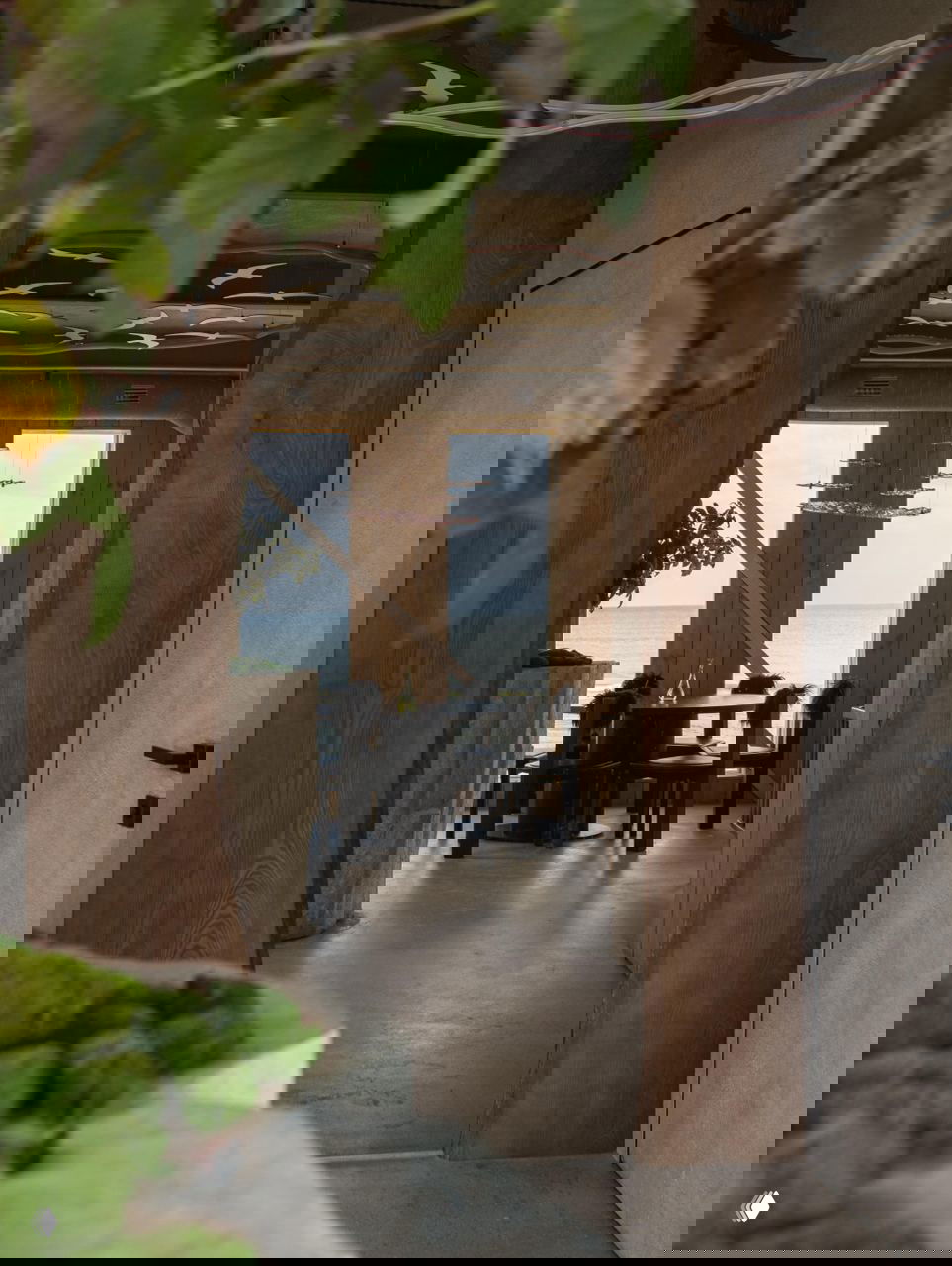 Проход через деревянную арку к столу у окна и виду на море: композиция из натурального дерева, зелёных растений и морского горизонта в кадре.