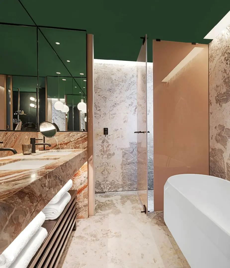 Ванная комната отеля: мраморная столешница и стеновые панели, стеклянная дверь душа и свободностоящая ванна на светлом мраморе.