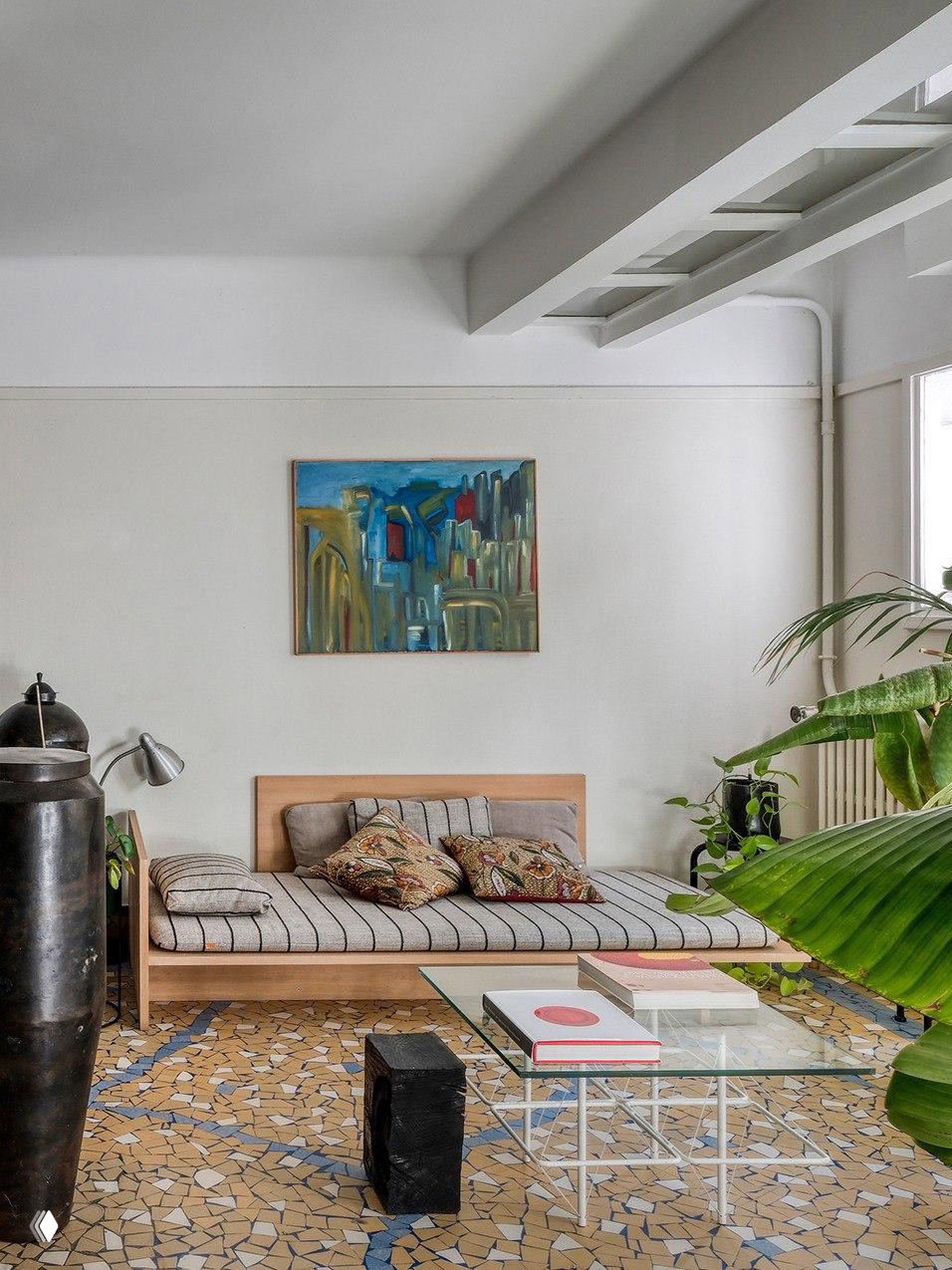 Спальная или гостевая зона с низкой деревянной кушеткой, абстрактной картиной на стене, зелёными растениями и мозаичным полом монастырского типа.