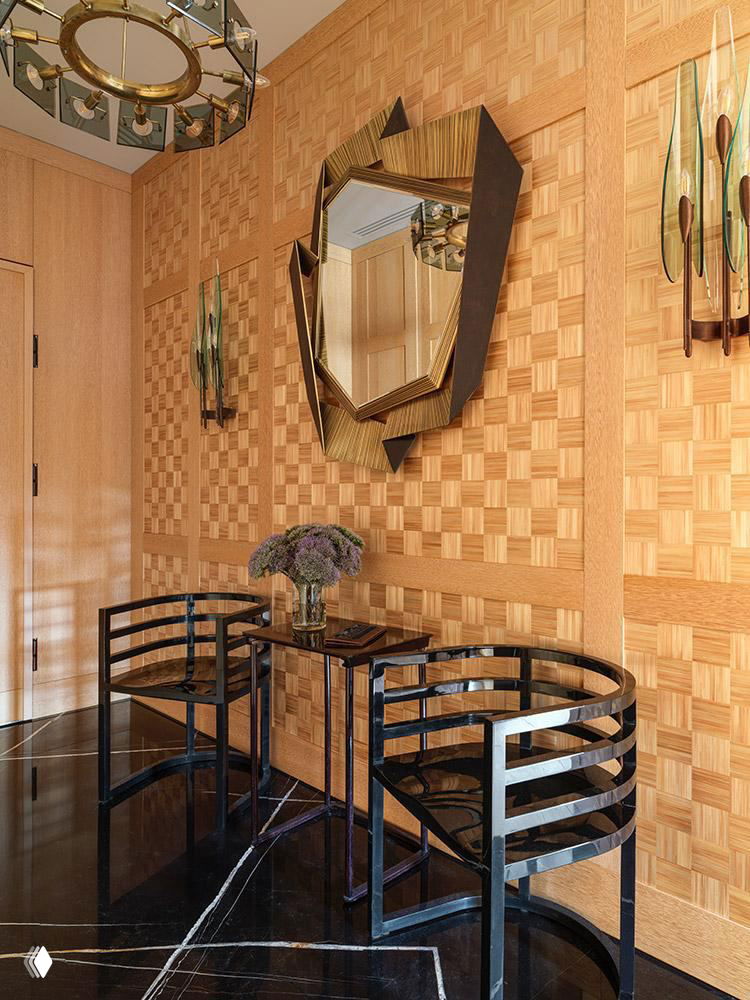 Прихожая с геометрической деревянной облицовкой стены, декоративным зеркалом неправильной формы и черными дизайнерскими стульями у консоли.