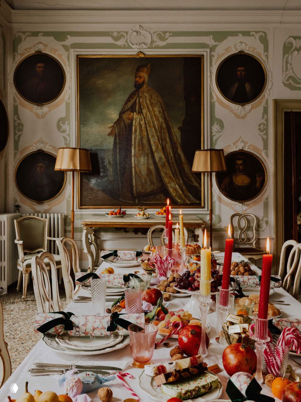 Большая столовая палаццо с фресками и круглыми портретами на стенах, сервированный праздничный стол с разноцветными свечами и десертами.