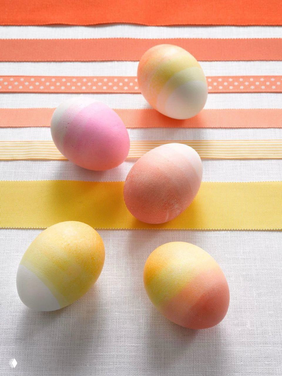 Пасхальные яйца в нежных пастельных оттенках — жёлтые, розовые, голубые — разложены на фоне разноцветных атласных лент, аккуратная декоративная композиция.