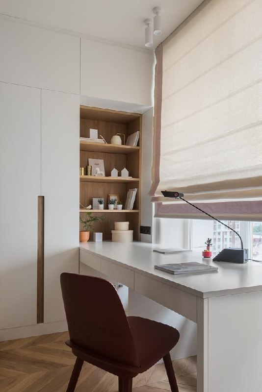 Рабочая зона у окна в московской квартире: белый письменный стол, встроенные открытые полки с аксессуарами и римская штора; функциональный стиль для деловых визитов.