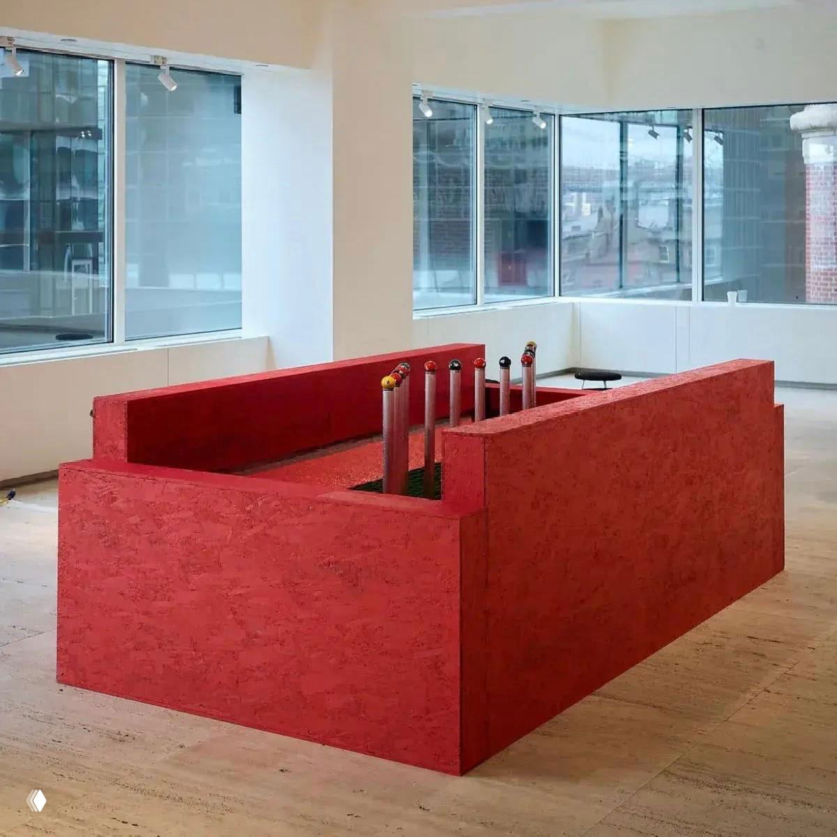 Современная галерейная инсталляция: красный прямоугольный подиум с вертикальными цилиндрическими элементами на фоне больших окон выставочного зала.