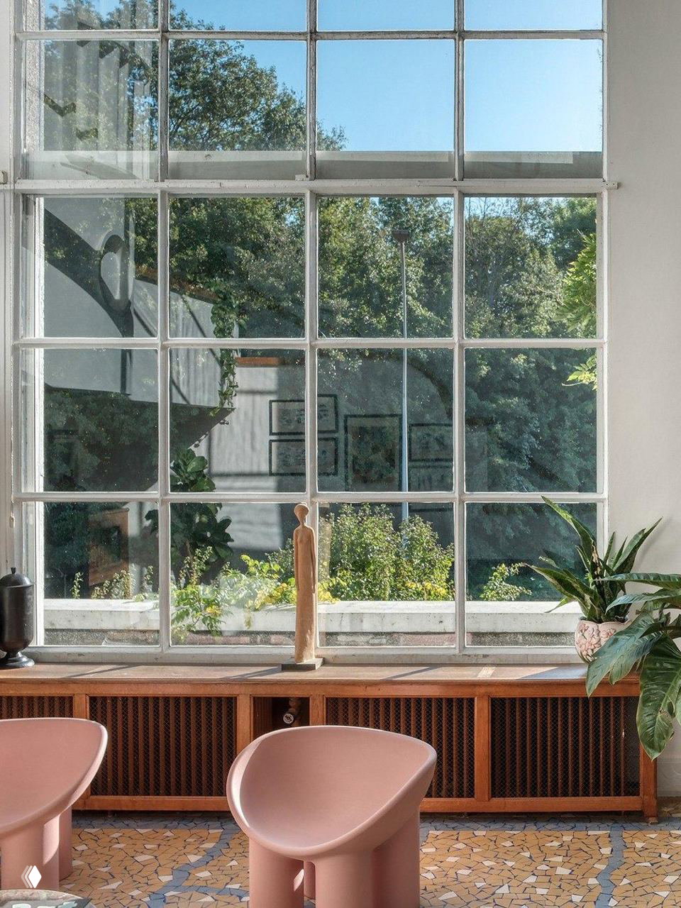 Большое остекление в виде квадратно‑рамного окна, через которое видна зелёная окружающая растительность; на переднем плане розовые кресла и мозаичный пол.