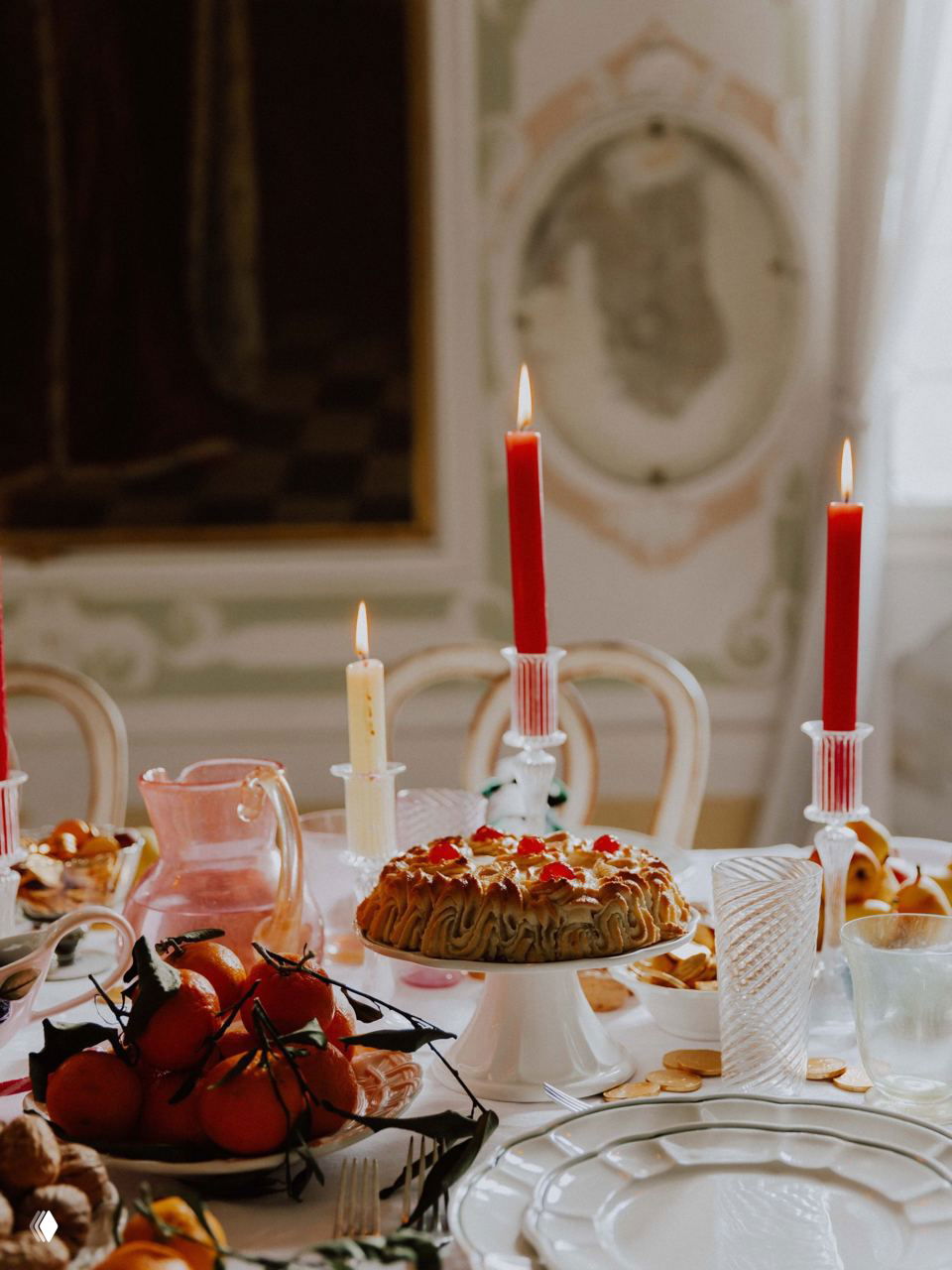 Крупный план праздничного стола: красные и кремовые свечи, пирог на ножке, фрукты и стеклянная посуда на фоне лепнины интерьера.
