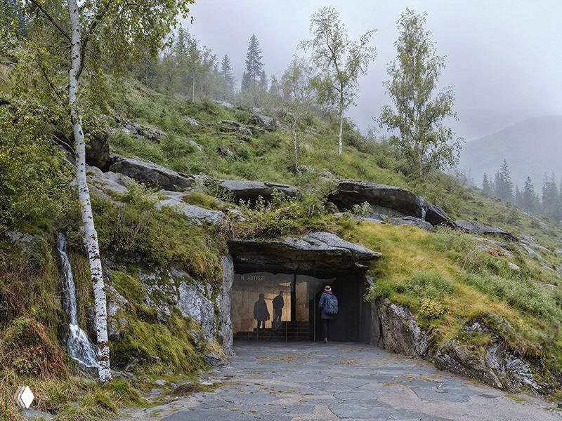 Вход в подземный музей The Cave, врезанный в скалистый склон у озера; видна узкая трещина-вход, силуэты людей и берёзы на склоне.