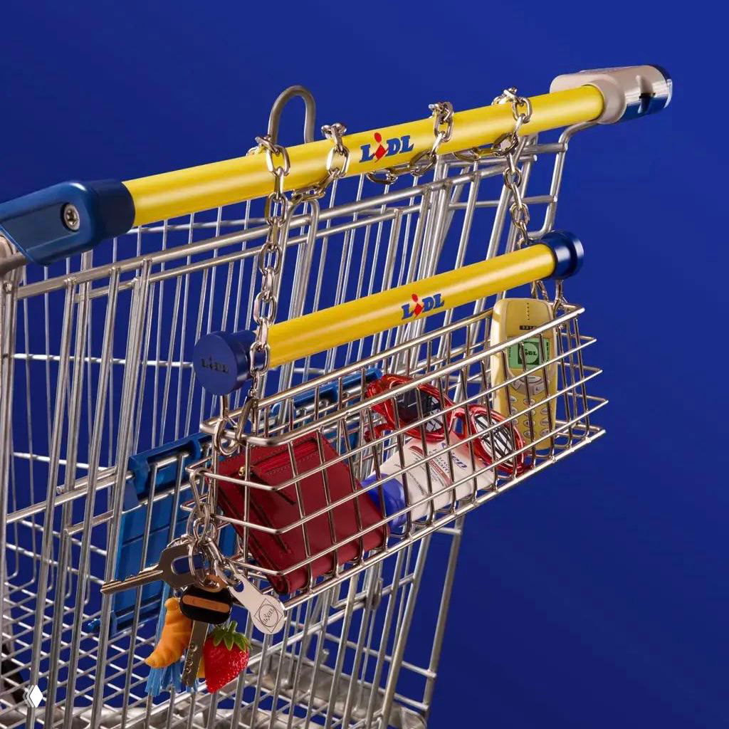 Крупный план подвесной корзины‑сумки на тележке с жёлтыми ручками и цепочками, видны брелоки и детали — акцент на промышленный дизайн коллаборации.