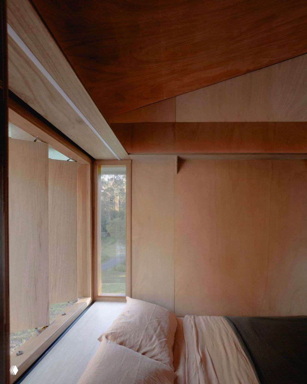 Угол спальни с крупным окном и ставнями: постель, деревянная отделка и мягкий дневной свет, подчёркивающий естественные материалы.