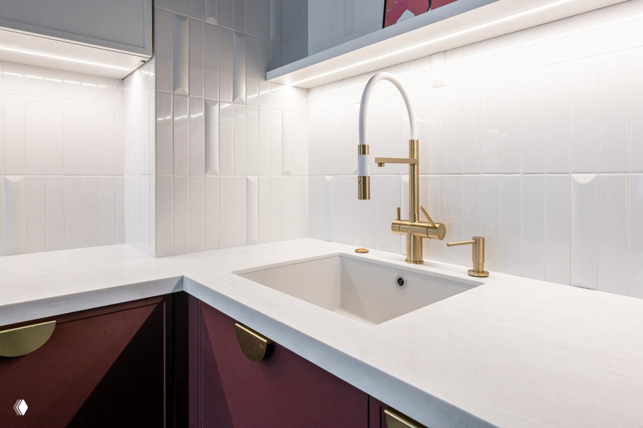 Крупный план зоны мойки в кухне «Броуди»: столешница из искусственного камня, подстольная мойка и выразительный золотой смеситель на светлом фоне плиточного фартука.