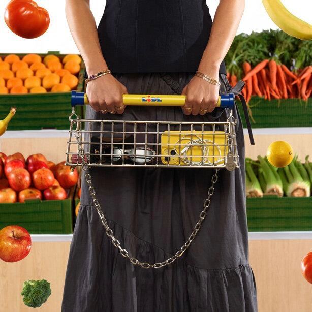 Человек держит сумку‑тележку в руках на фоне прилавка с овощами; показана форма корзины, пропорции сумки относительно тела и контекст использования.