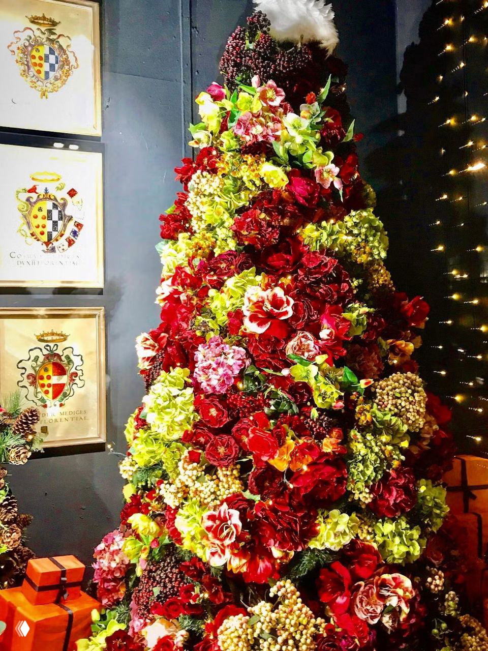 Плотно украшенная елка, покрытая крупными искусственными цветами и ягодными гирляндами, насыщенная красно‑бордовыми и зелеными оттенками в интерьере шоурума.
