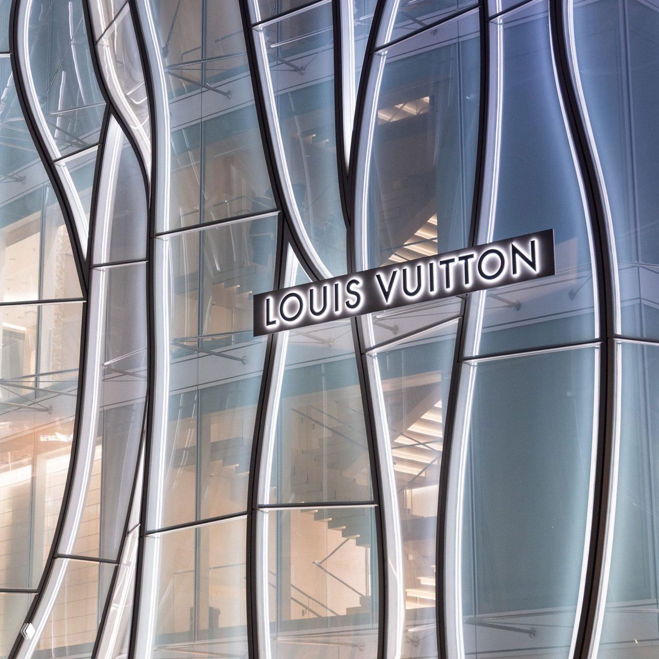 Крупный план фасадной конструкции Louis Vuitton: изогнутые стеклянные панели с тонкими металлокаркасами и неоновой подсветкой вокруг логотипа.
