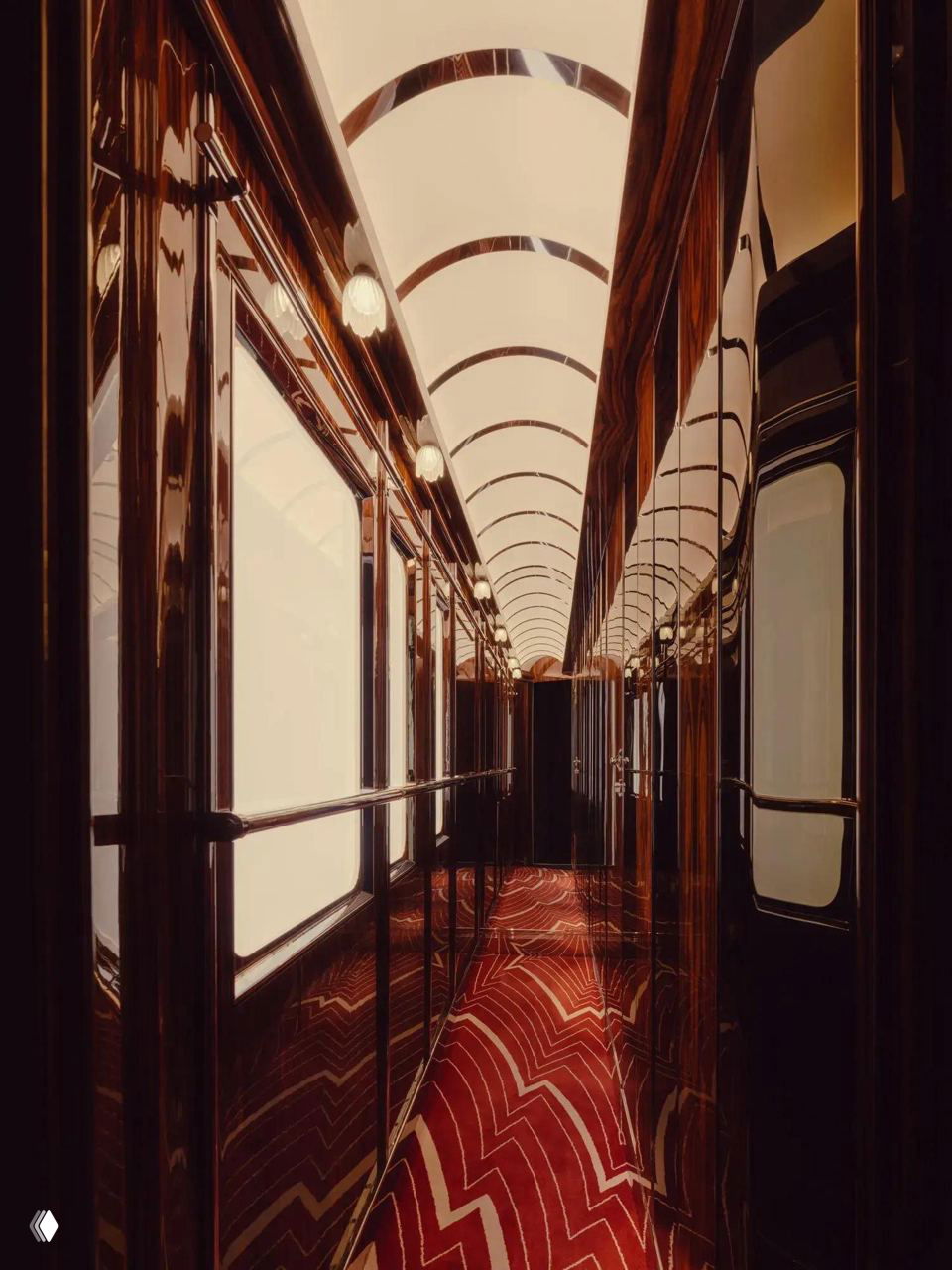 Узкий коридор вагона с полированными стенами, волнообразным освещением потолка и красным ковровым покрытием с геометрическим узором ар‑деко.