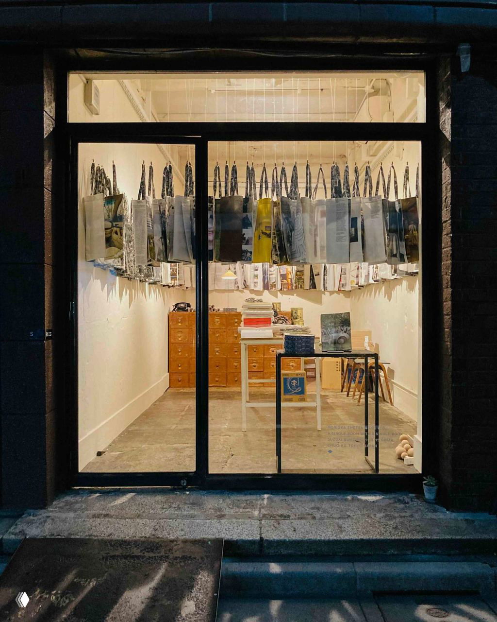 Окно магазина: в центре один стол, по стенам и сверху подвешены листы и страницы, вокруг — аккуратная деревянная мебель и минималистская витрина.