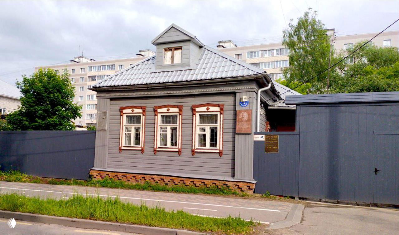 Небольшой деревянный дом с резными наличниками и мемориальной табличкой у стены, зелёная трава и тротуар на фоне многоэтажек — городской уголок Клину.