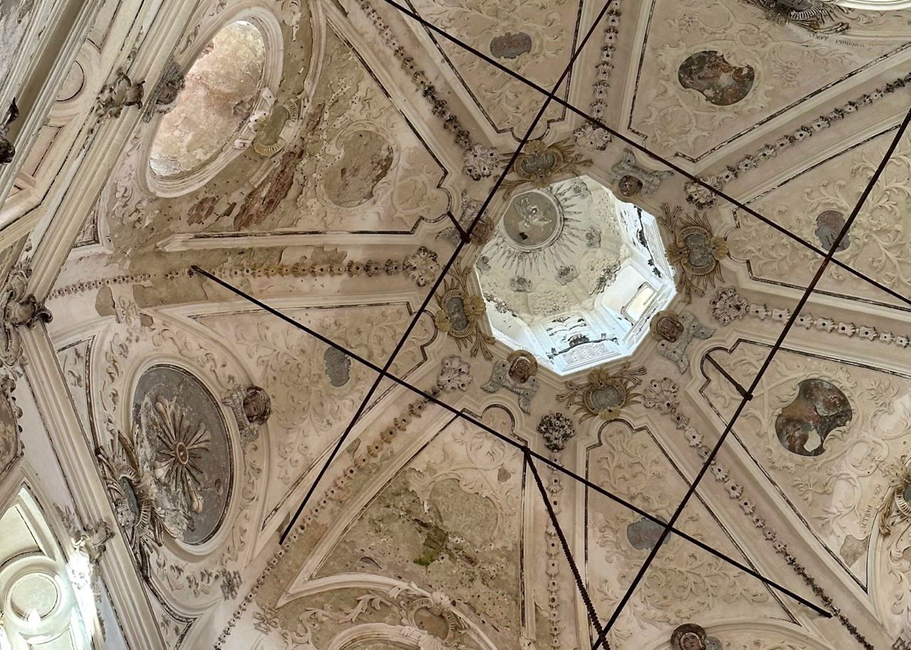 Декор потолка и купола с барочной лепниной и фрагментами фресок, видны поддерживающие тросы и участки разрушений, требующие консервации.