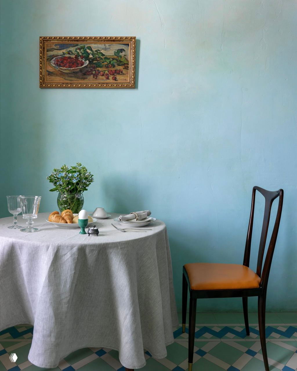 Небольшая столовая с голубой стеной и круглым столом; на полу плитка в клетку зелёно‑голубых оттенков создаёт лёгкую винтажную атмосферу.