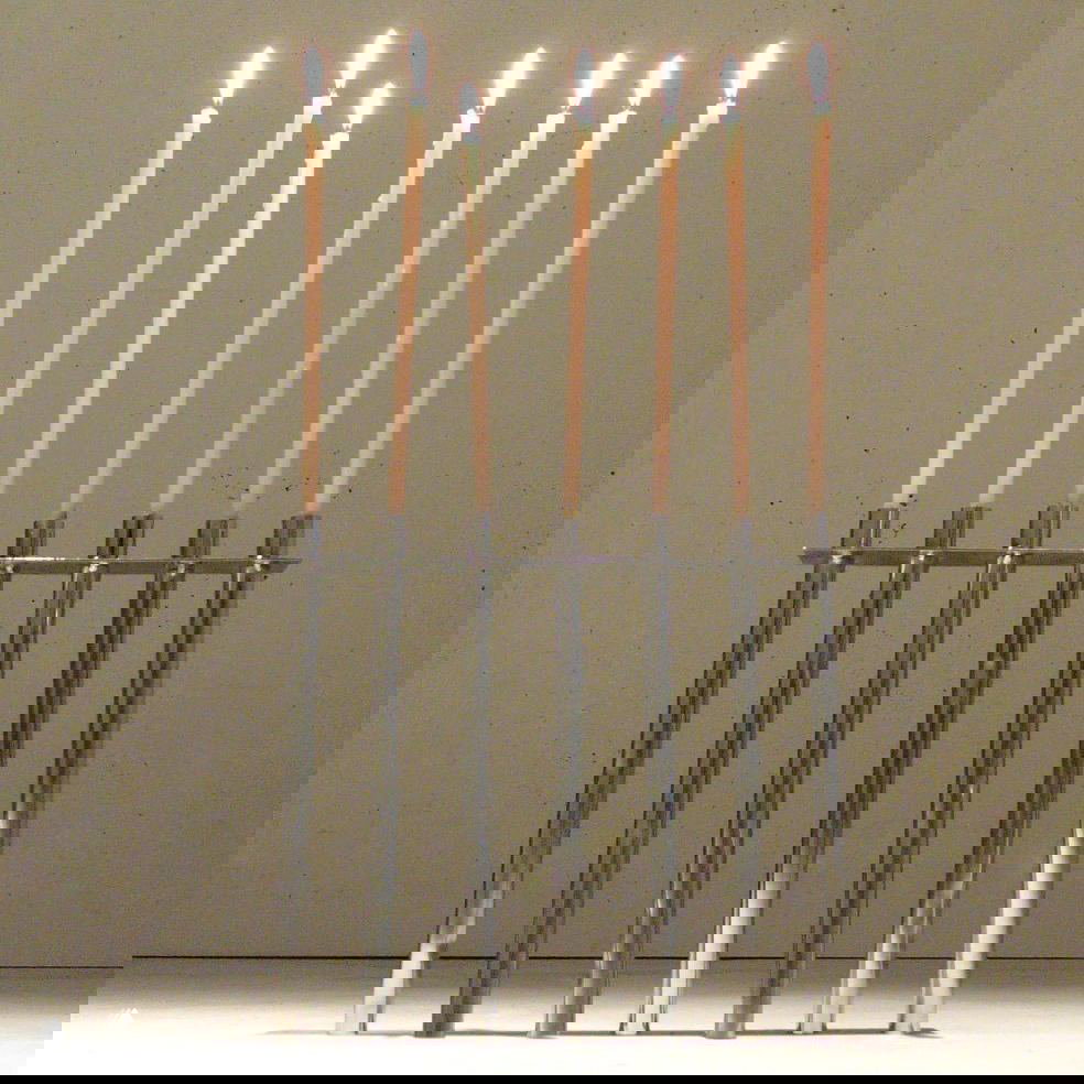 Высокий подсвечник из полированных стальных стержней: семь тонких свечей установлены в минималистскую конструкцию на нейтральном фоне.