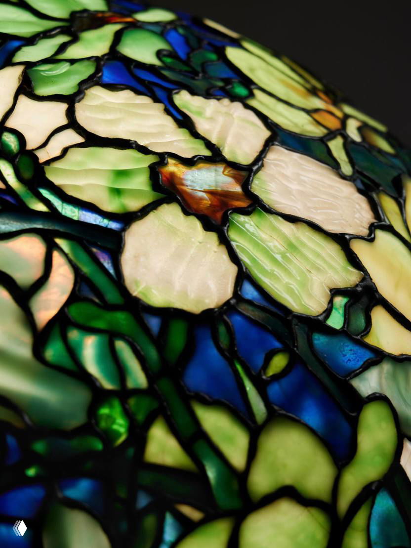 Крупный план витражного абажура Tiffany «Magnolia»: перламутровые лепестки магнолии, синие и зелёные стекла и тёмные припои в деталях.