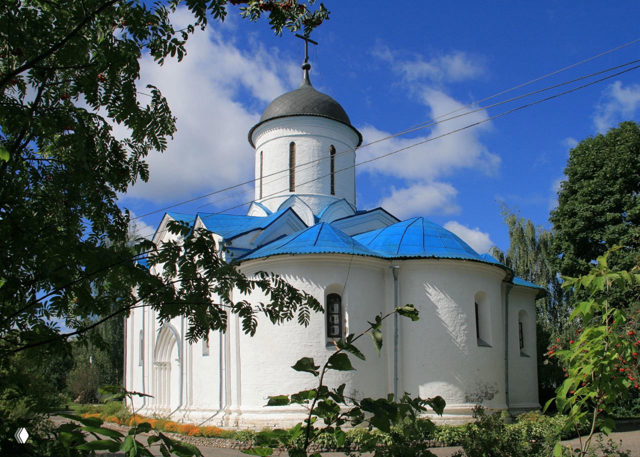 Белая старинная церковь с круглой апсидой и куполом, ярко-синие крыши и крест на фоне голубого неба и зелёных деревьев в летний день.