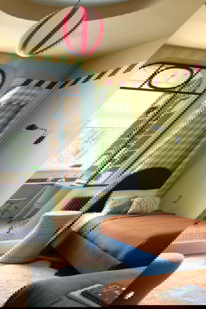 Одна из кроватей с зелёным клетчатым балдахином, оранжевым покрывалом и плетёным пуфом рядом; на заднем плане большой рабочий стол у окна.