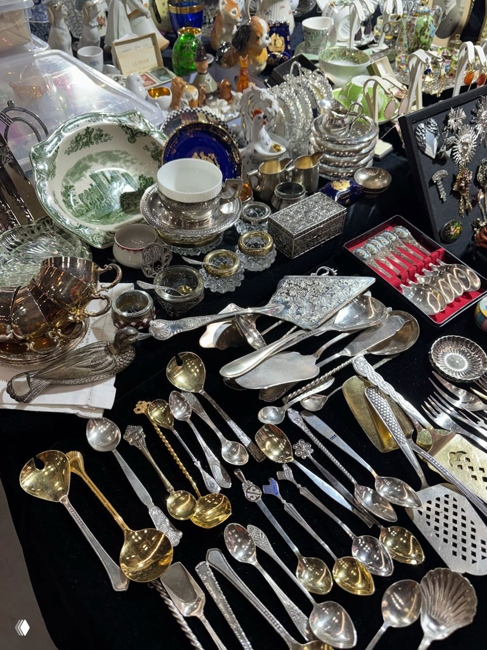 Стол с большим количеством серебряных приборов, ложек и сервировочных предметов, разложенных для продажи на блошином рынке.