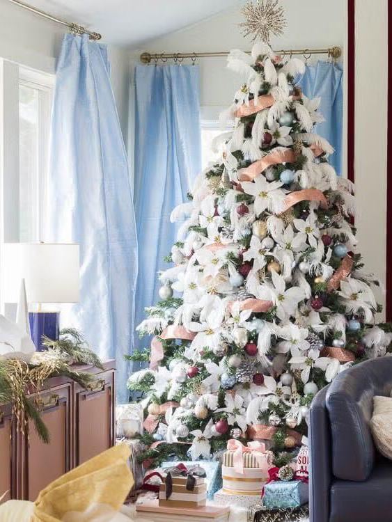 Тонкая высокая елка в светлом помещении, украшенная пастельными помпонами и подставкой с подарками, создающая мягкий и игривый праздничный образ интерьера.