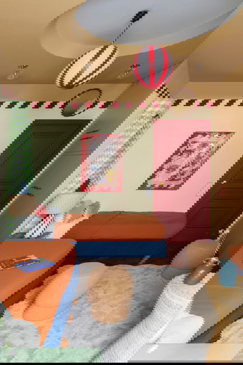 Две односпальные кровати с тёмно‑синими основаниями и оранжевыми покрывалами, красная дверь и подвесной светильник в центре потолка, декор на стене.