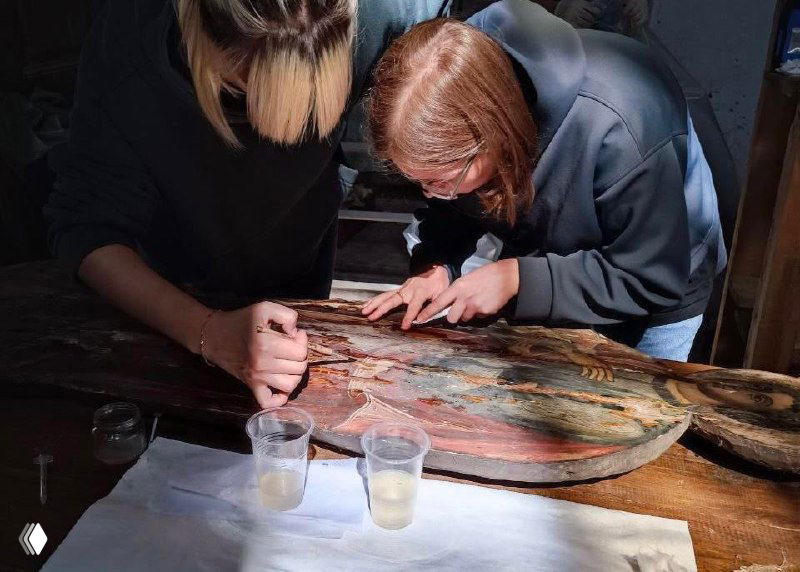 Реставраторы за работой: две женщины аккуратно очищают иконостенную панель на столе в мастерской, рядом инструменты и банки с растворами для консервации.