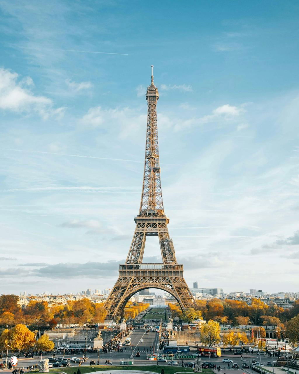 Классический вид на Эйфелеву башню на фоне голубого неба: стальная решётчатая структура и окружающие парки с людьми у основания монумента.